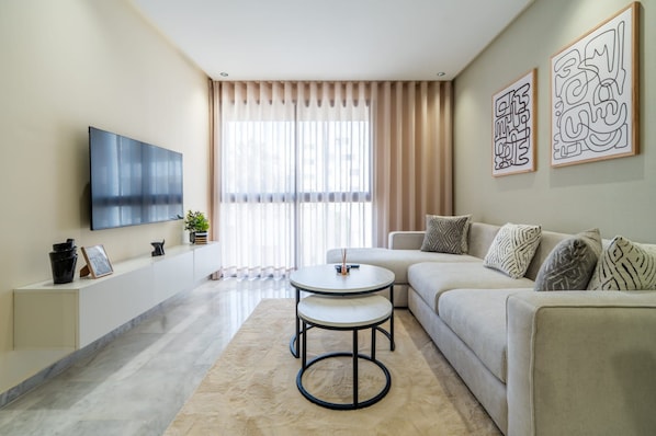 A modern living room with a flat-screen TV, a round coffee table, a sofa, and a large window with curtains.