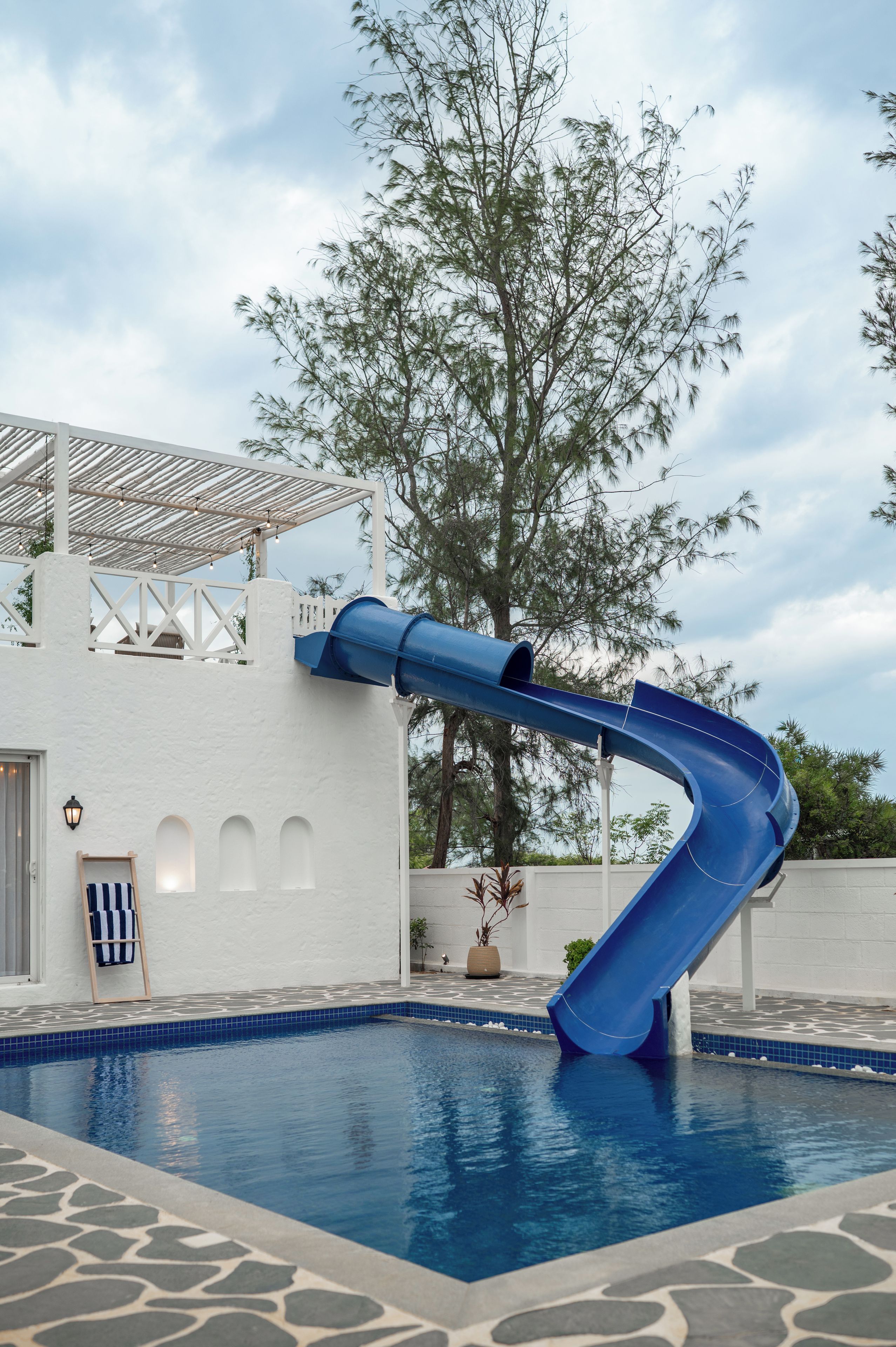 Una piscina con uno scivolo blu, un edificio bianco con finestre ad arco e un albero sullo sfondo.