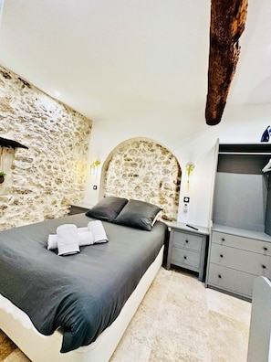 A bedroom with a stone wall, a bed with grey bedding, a nightstand, a dresser, and a wooden beam. - 16097522 FRGK 5-Apartment Building - Downtown Cannes - Conference Special - 47 rue Meynadier (Cannes)