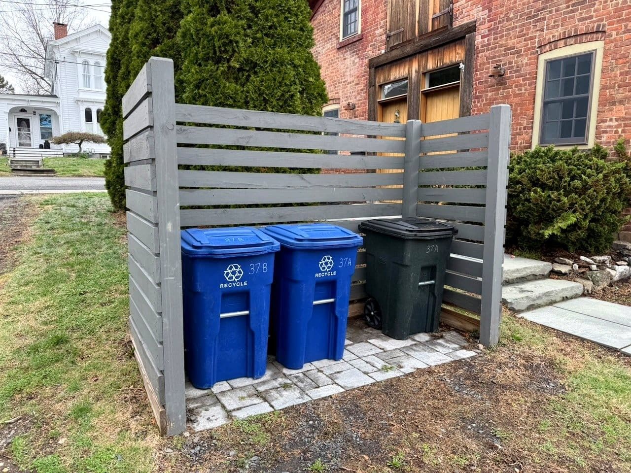 A grey fence with recycling bins, a brick building, and a well-maintained lawn.