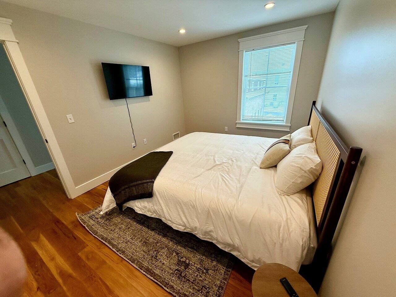 A bedroom with a bed, a television mounted on the wall, a window with blinds, and a wooden floor.