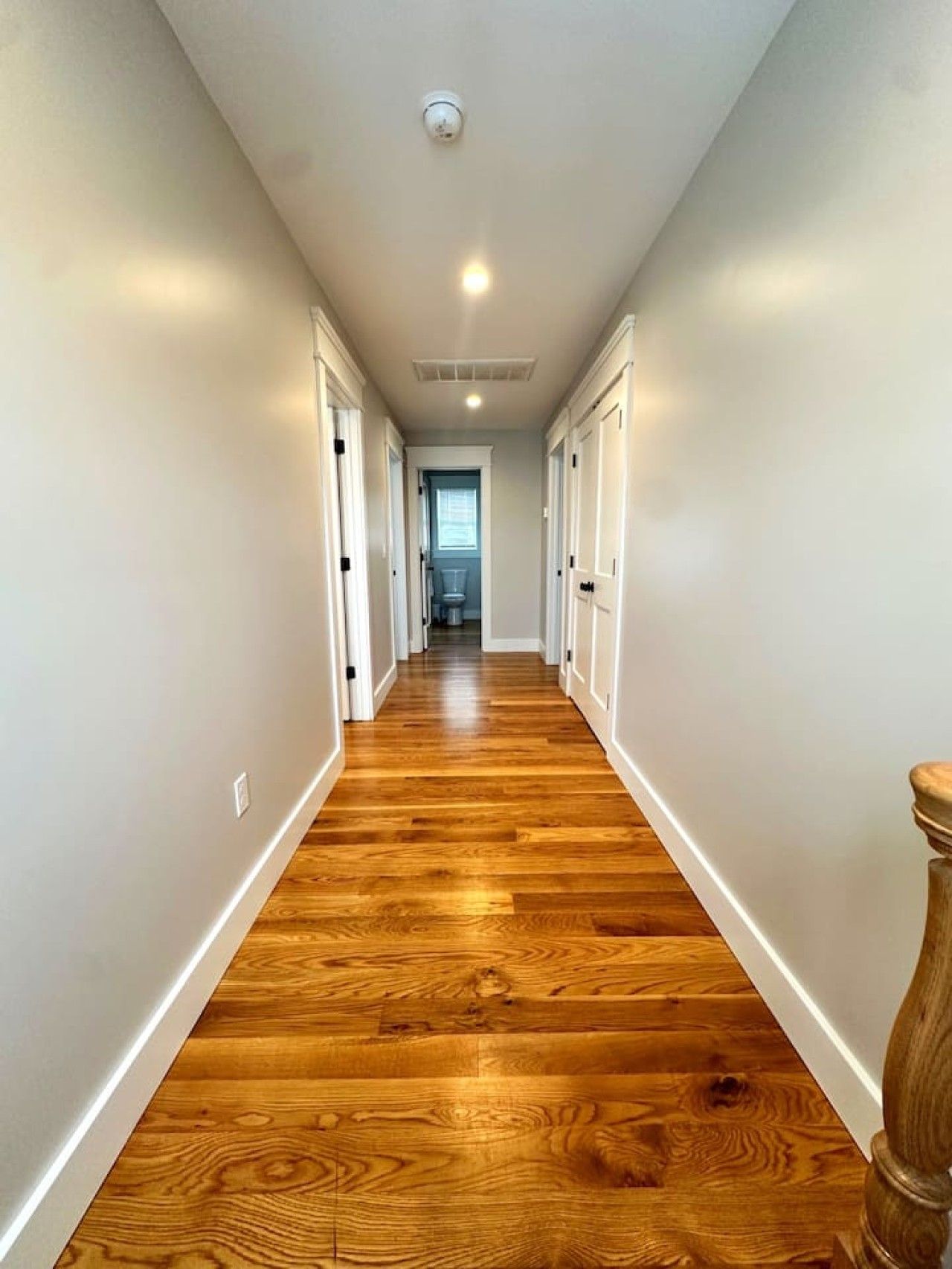 A hallway with wooden flooring and white walls, leading to a room with a window.