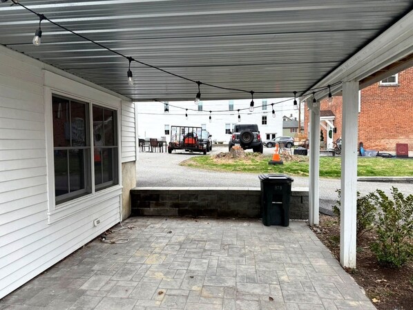 A covered outdoor area with a paved walkway, a trash bin, and a vehicle parked in the background.