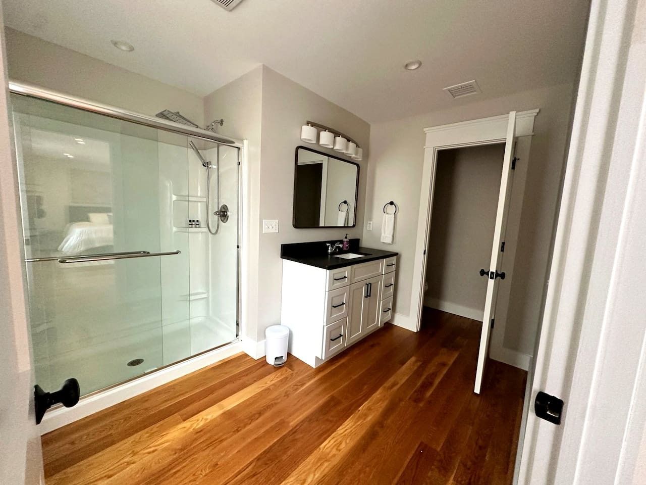 A modern bathroom with a glass shower enclosure, a white vanity with a black countertop, and a wooden floor.