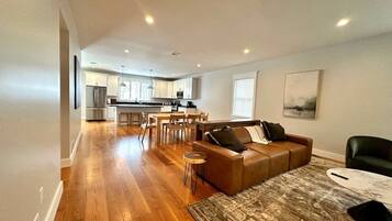 A spacious living area with a wooden floor, a leather sofa, a coffee table, and a dining area with chairs and a kitchen in the background.