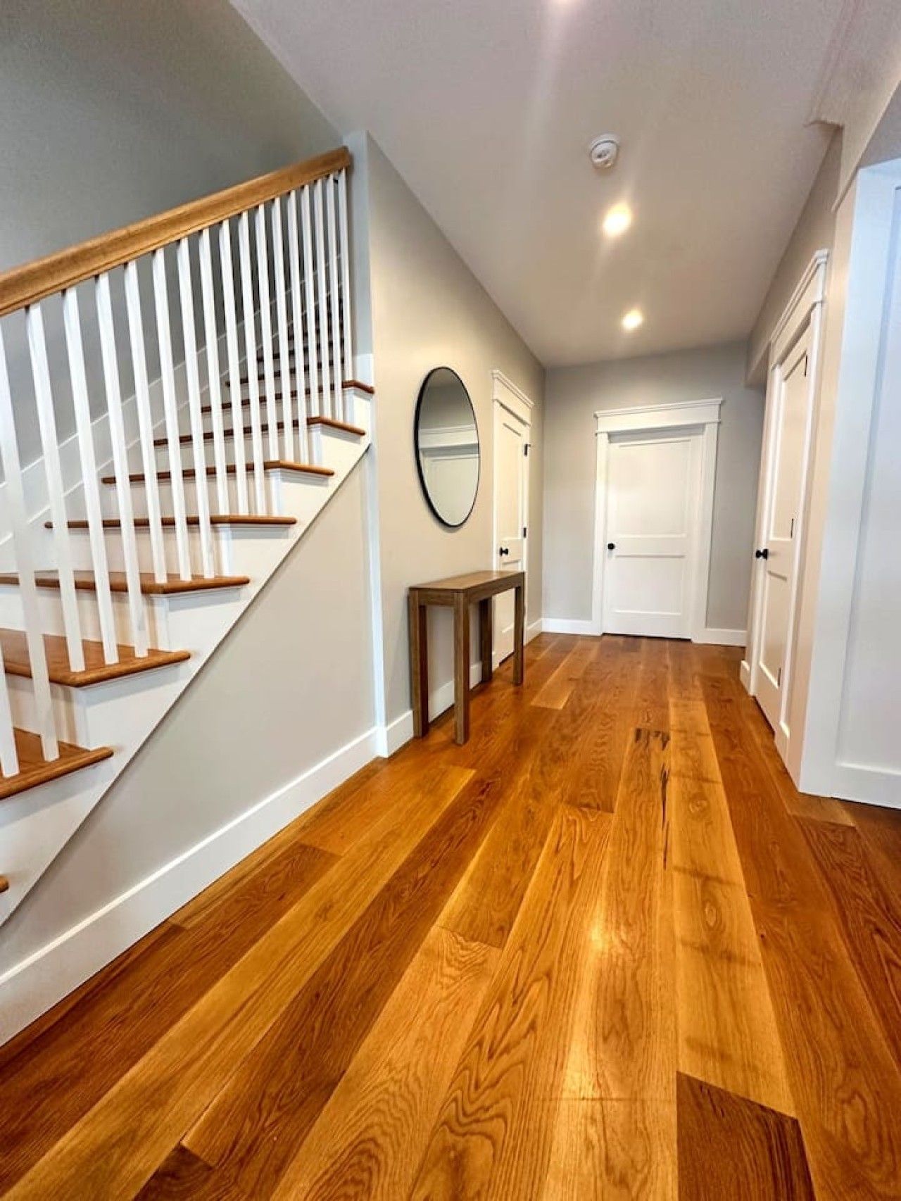 A hallway with wooden flooring, white walls, a staircase with white railings, and a door at the end.