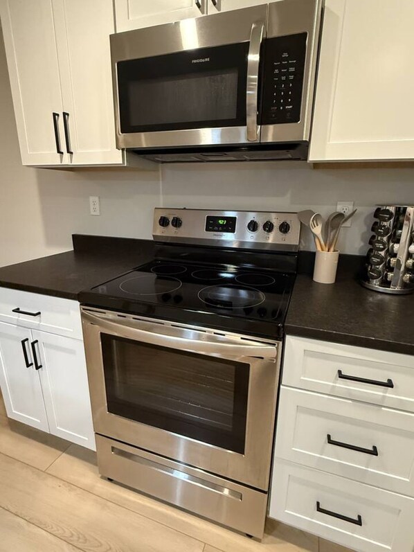 Una cocina con electrodomésticos de acero inoxidable, incluyendo un microondas y un horno, armarios blancos y encimera negra.