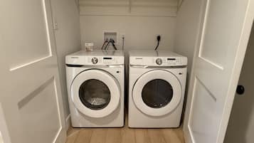 A laundry room with two white front-loading washing machines, a shelf above them, and a power outlet on the wall.