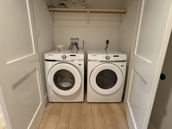 A laundry room with two white front-loading washing machines, a shelf above them, and a power outlet on the wall.