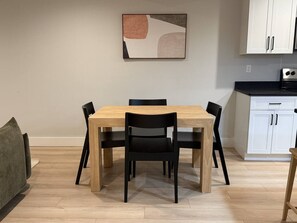 A dining area with a wooden table and chairs, a painting on the wall, and a kitchen with white cabinets.