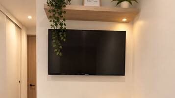 A dining area with a white table, two chairs, a TV mounted on the wall, and a shelf with plants and a framed quote.