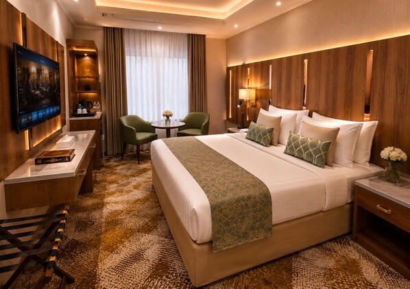 A hotel room with a large bed, a desk with a flat-screen TV, a small table with chairs, and a view of the outdoors through the window. - HINDIBAA HOTEL MAKKAH (Makkah)