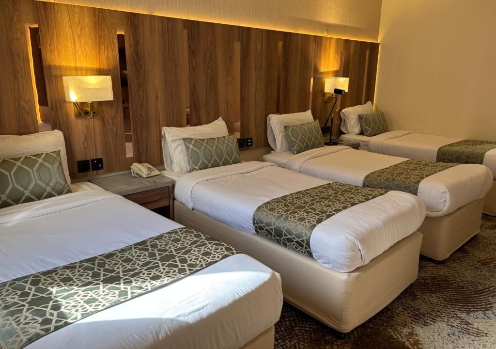 Habitación de hotel con cuatro camas dispuestas en fila, cada una con almohada y mesita de noche.