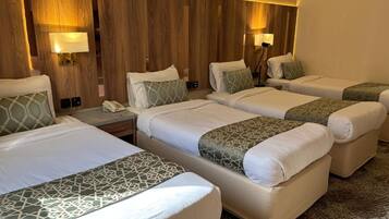 Habitación de hotel con cuatro camas dispuestas en fila, cada una con almohada y mesita de noche.
