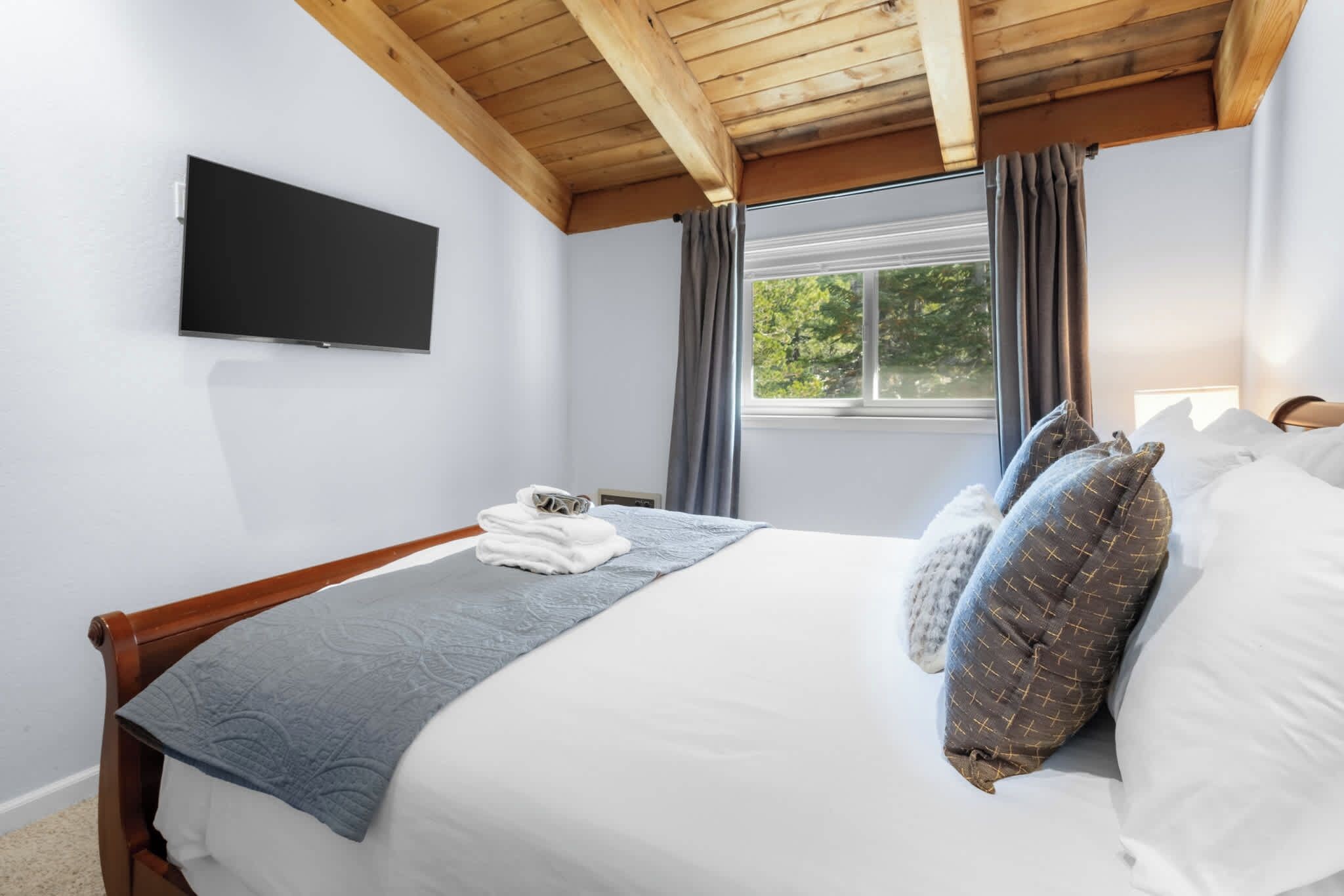 A bedroom with a bed, a flat-screen TV, a window with curtains, and a wooden ceiling.