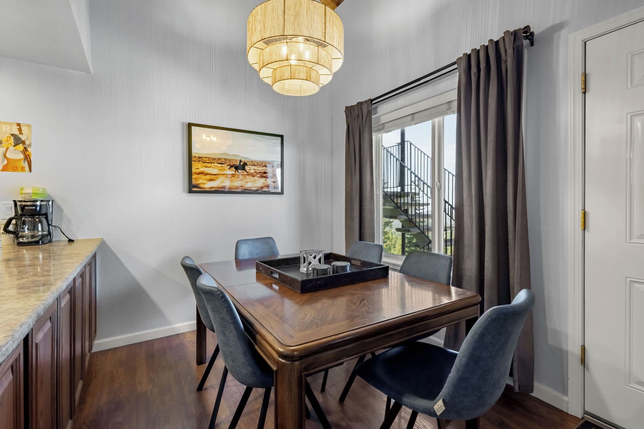 A dining area with a wooden table, six chairs, a coffee maker, a framed picture, and a window with curtains.