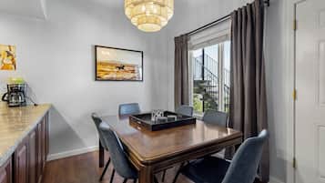 A dining area with a wooden table, six chairs, a coffee maker, a framed picture, and a window with curtains.