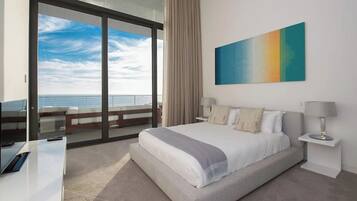 A hotel room with a large bed, a desk, a chair, a nightstand, a lamp, a painting, and a view of the ocean.