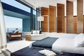 Ett modernt sovrum med en stor säng, en sittgrupp med en soffa och ett sidobord, och utsikt över havet.