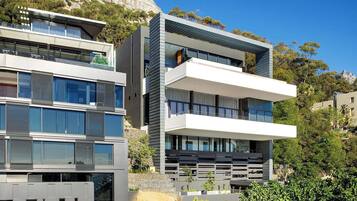 A multi-story modern building with balconies, large windows, and a rocky hillside background.