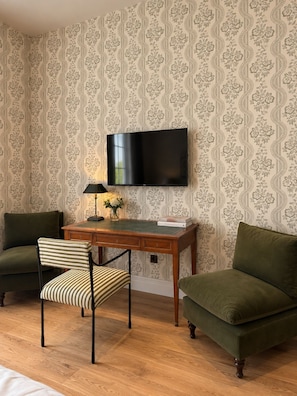 Een kamer met een flatscreen televisie aan de muur, een houten bureau met een lamp, een stoel en een bank.