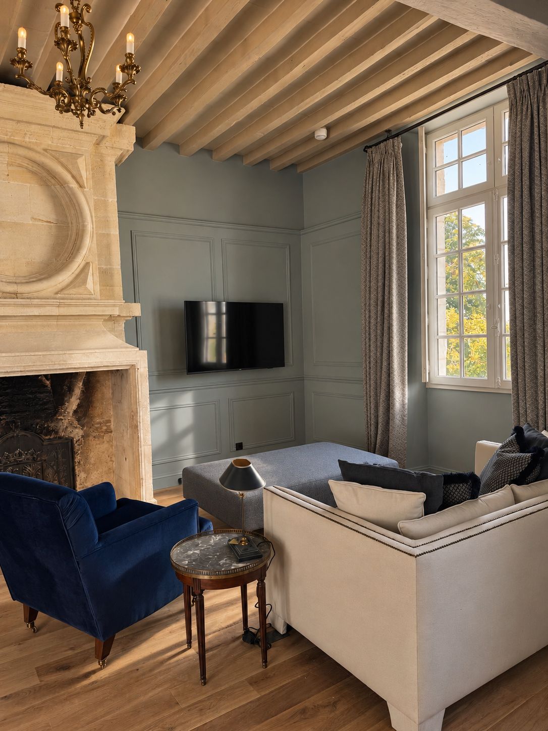 Una sala de estar con chimenea, televisor montado en la pared, un sofá, un sillón azul y una pequeña mesa redonda.
