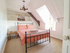 Ein Schlafzimmer mit schrägem Dach, einem Bett mit kariertem Überwurf, einem Nachttisch mit Lampe und einem Bücherregal voller Bücher.
