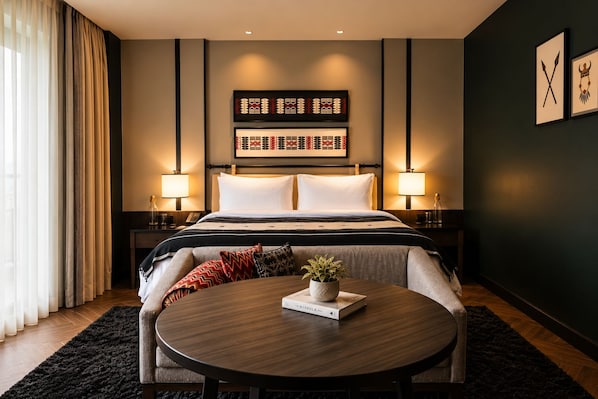 A hotel room with a bed, bedside lamps, a round table with a book, a potted plant, and a dark-colored wall with framed artwork.