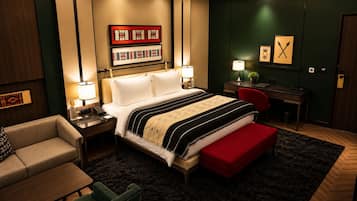 A hotel room with a bed, bedside tables, a desk, a chair, a sofa, and artwork on the walls.