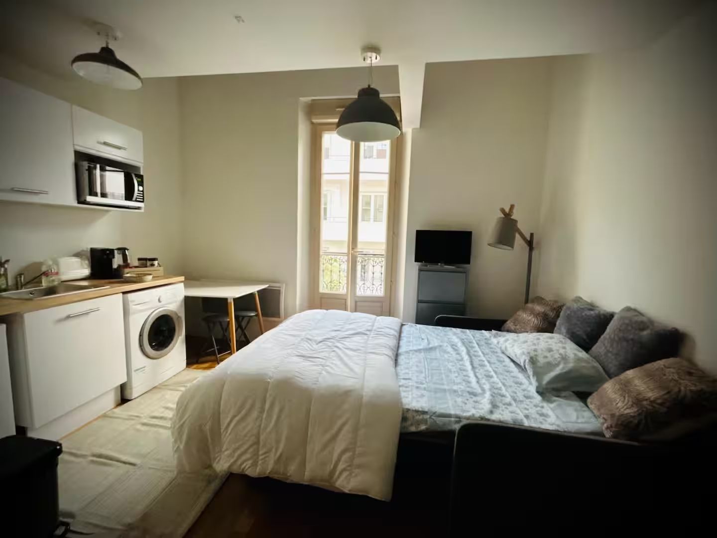 A bedroom with a bed, a desk, a washing machine, a television, and a balcony.