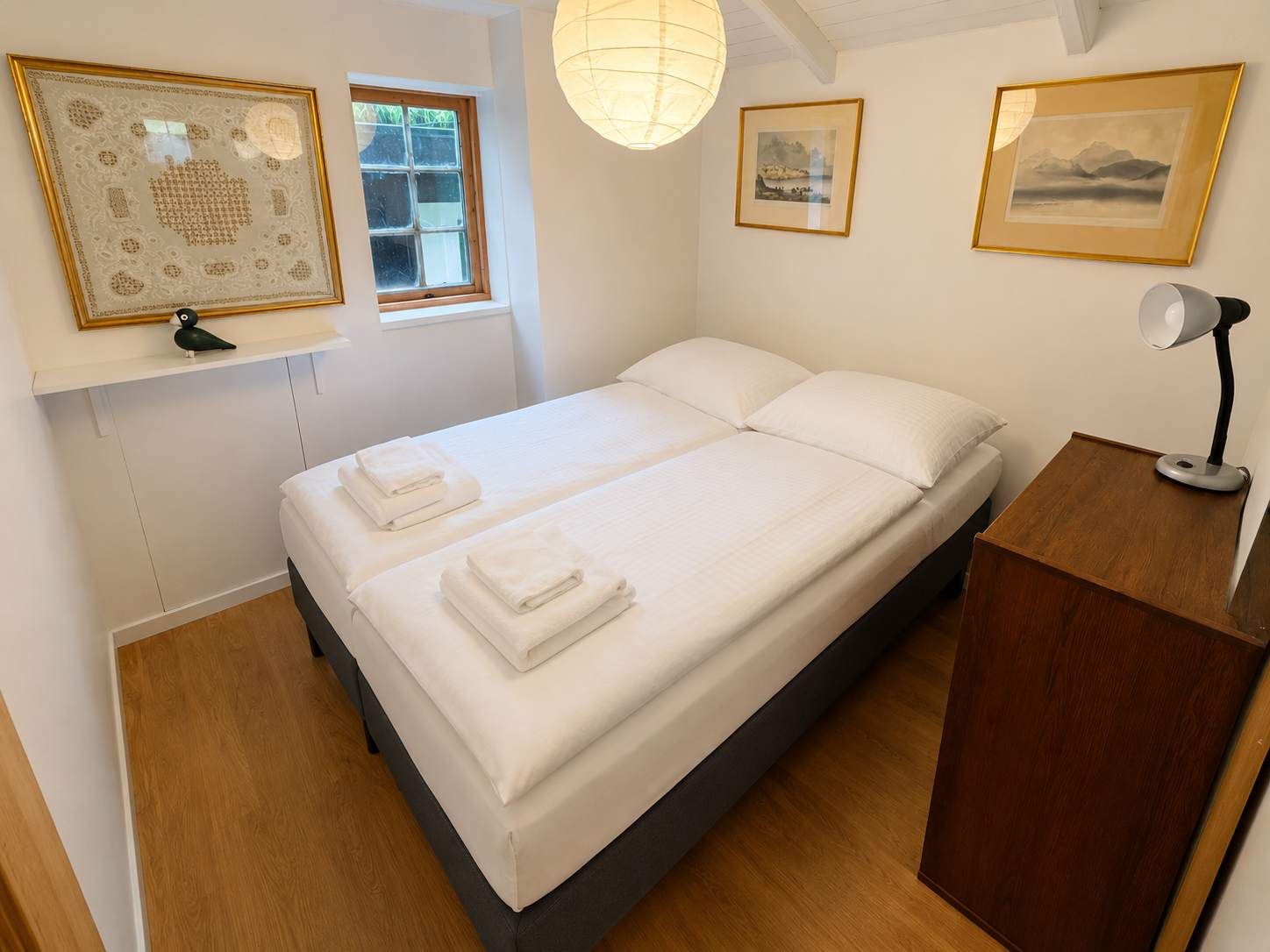 Uma cama de casal com lençóis brancos, uma mesa de cabeceira com abajur e quadros nas paredes.