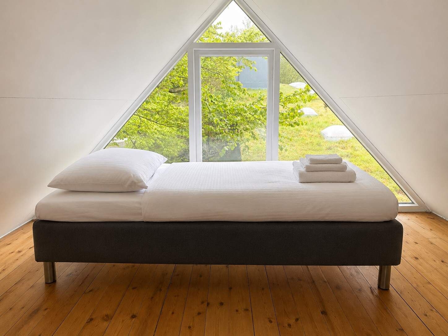 A bed in a triangular room with a large window, wooden flooring, and a view of greenery outside.