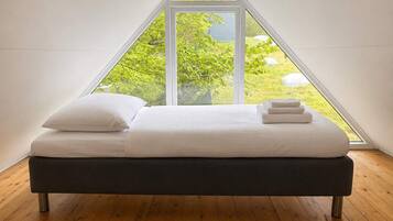 A bed in a triangular room with a large window, wooden flooring, and a view of greenery outside.