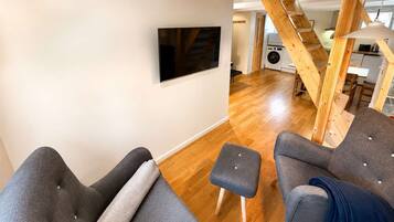A modern living room with a flat-screen TV mounted on the wall, grey upholstered sofas, a small ottoman, and a wooden staircase leading to an upper level.
