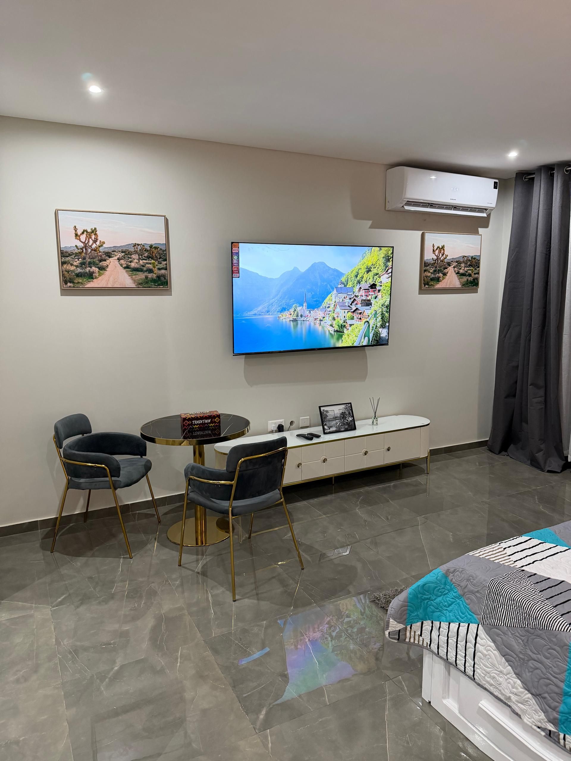 A modern living room with a flat-screen TV, two chairs, a round table, a white cabinet, and a bed with a patterned blanket.