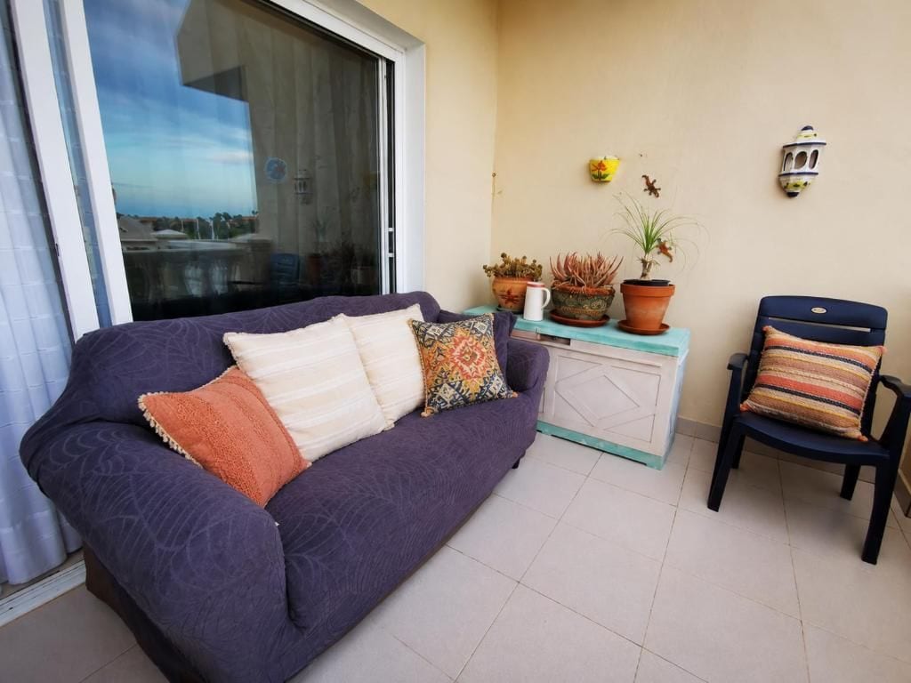 Una habitación con un sofá morado, una mesita con plantas en macetas, una silla y un ventanal corredera.