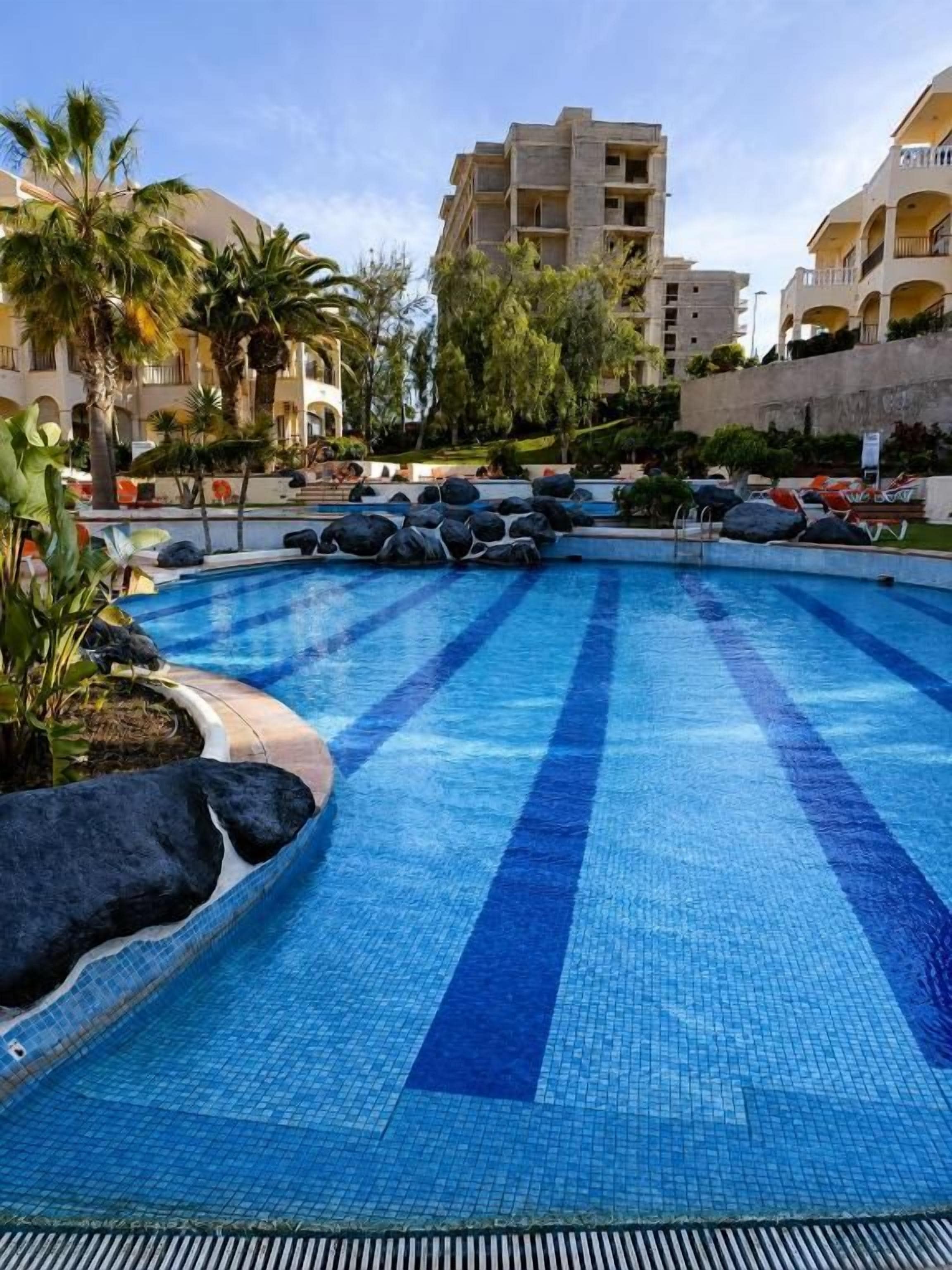 Una piscina con agua azul cristalina, rodeada de rocas y plantas, con un edificio al fondo.