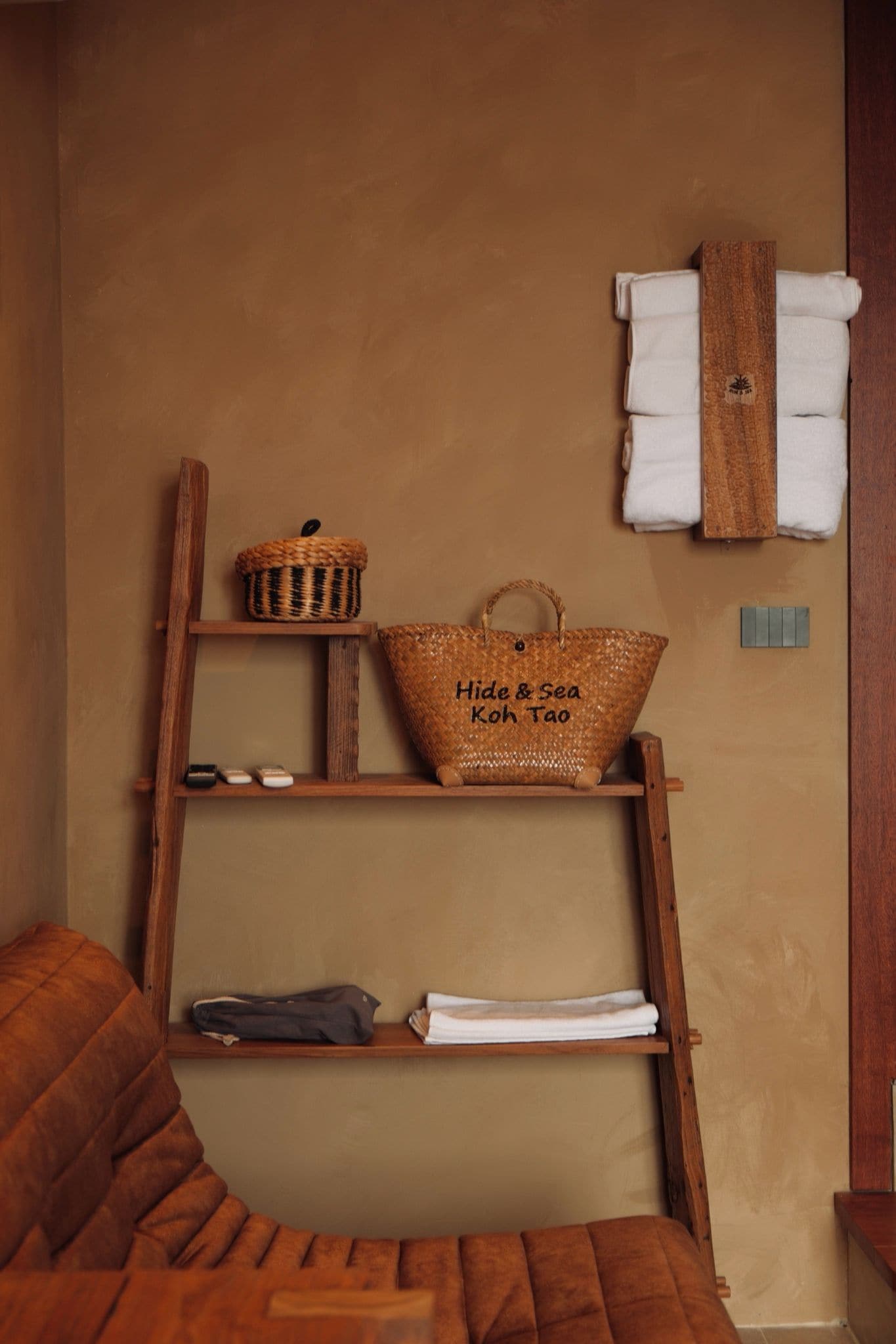 A wooden ladder shelf with a woven basket, a tote bag, and folded towels against a beige wall.