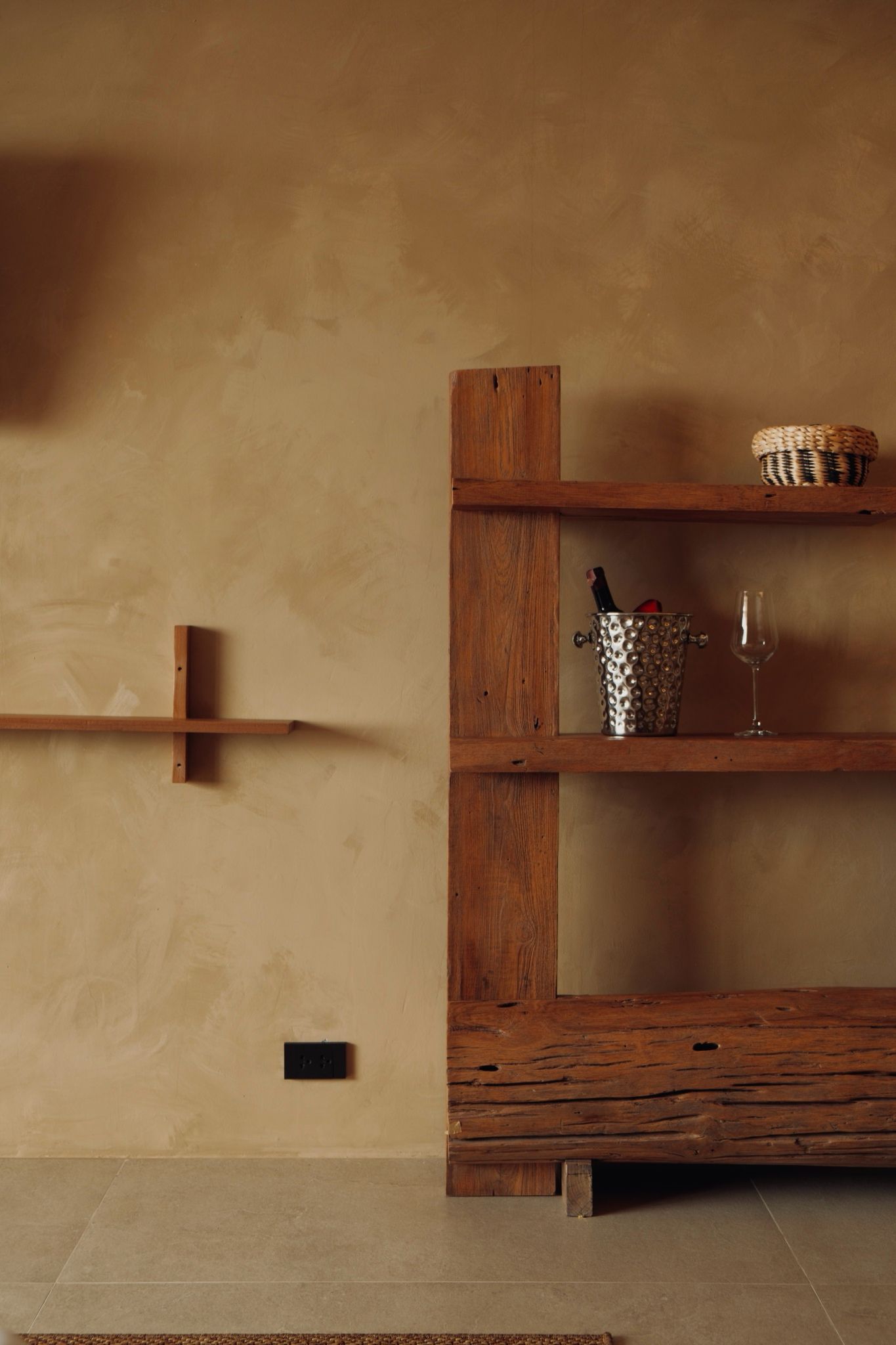 Un estante de madera con una botella de vino y una copa, una cesta tejida y un enchufe en la pared.