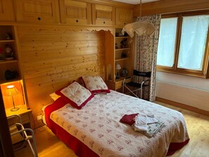 A bedroom with a wooden headboard, a bed with a patterned bedspread, a bedside table with a lamp, a chair, and a window with curtains. - Beautiful one-bedroom Cocoon apartment with a view of the Dents du Midi (Ollon)