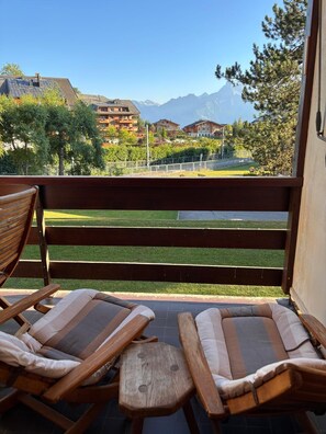 A balcony with two wooden lounge chairs and a small table, overlooking a well-maintained lawn and a mountainous landscape. - Beautiful one-bedroom Cocoon apartment with a view of the Dents du Midi (Ollon)