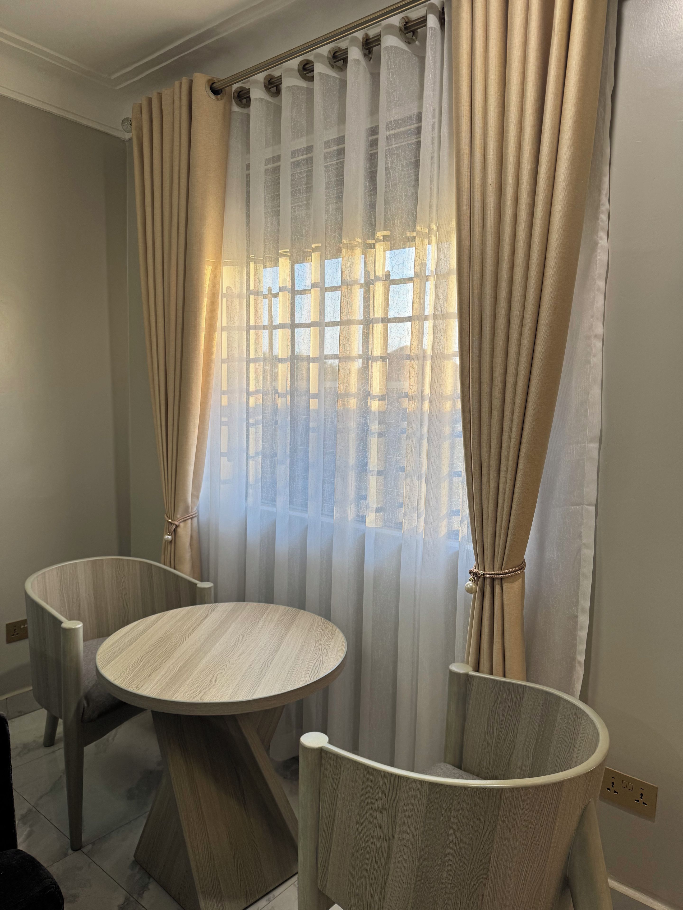 A room with a round table and two chairs, a window with curtains, and a wall socket.