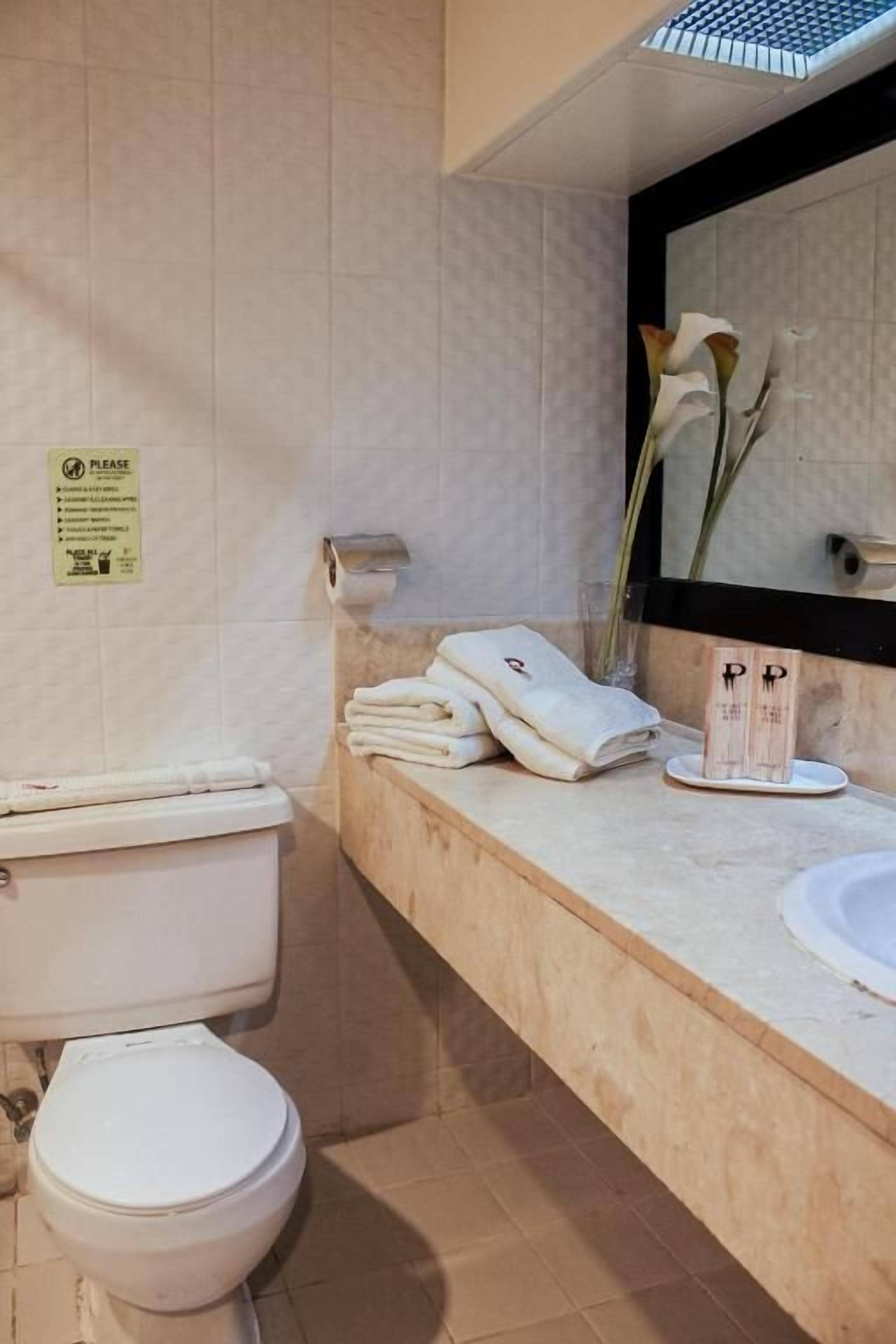 Un baño con inodoro, lavamanos y toallas. Hay un cartel en la pared y un florero con flores.