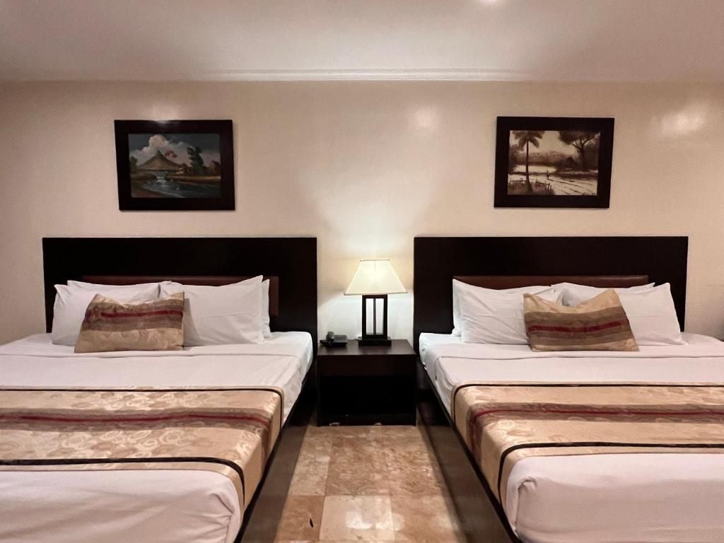 Habitación de hotel con dos camas, una mesita de noche con lámpara y dos cuadros en la pared.