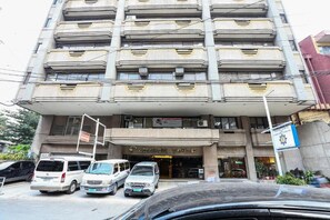 Un hotel de varios pisos con una entrada destacada, varios vehículos estacionados y un letrero con el nombre de un negocio.