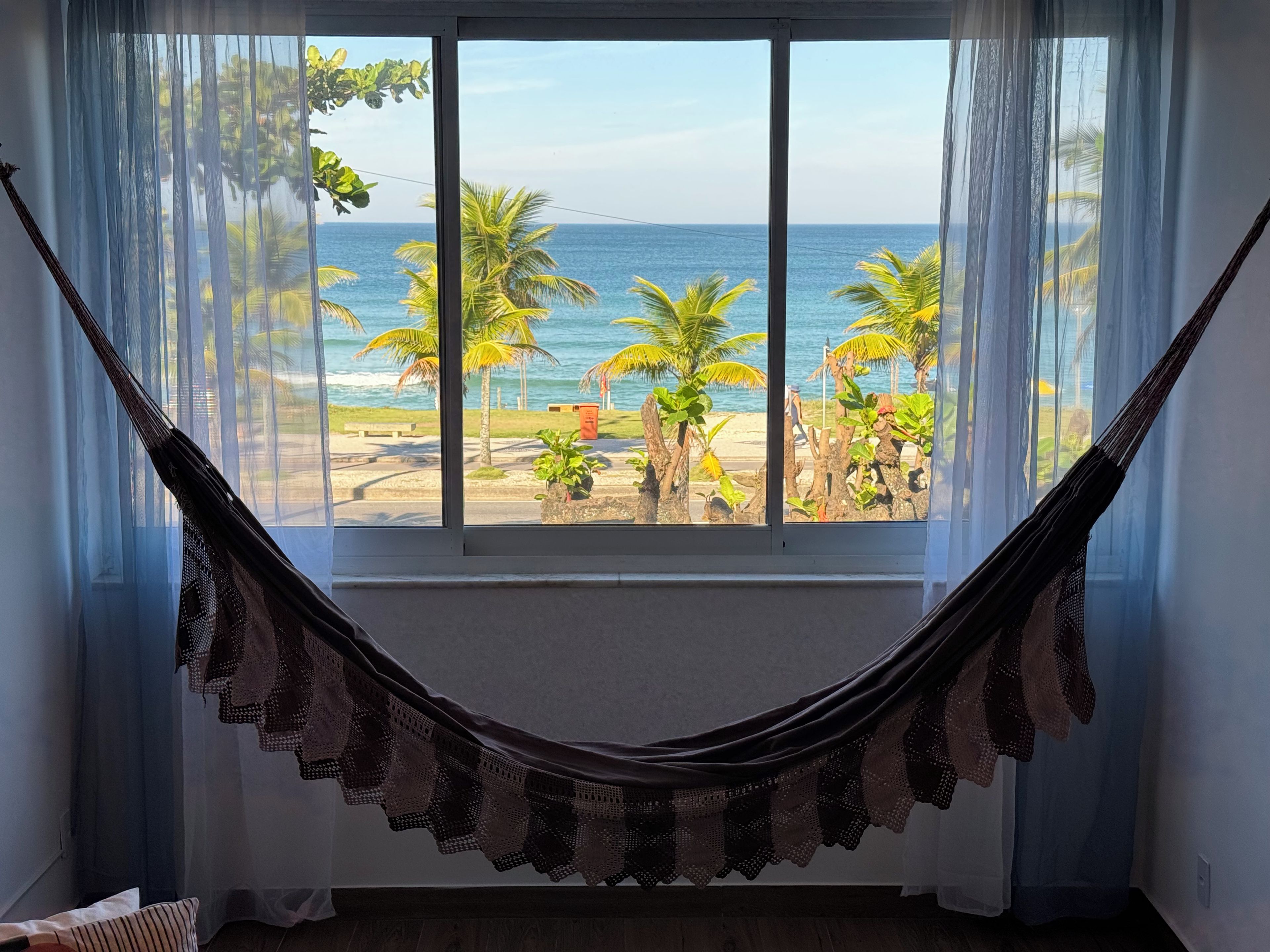 A hammock tied to a window with a view of a beach and palm trees.
