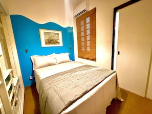 Ein Schlafzimmer mit einem Bett, einem Fenster mit Jalousien, einer Wand-Klimaanlage und einem gerahmten Bild.
