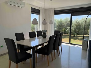 A modern dining area with a white table and black chairs, large windows with a view of greenery, and pendant lighting. - Holiday Home 'Aurora' with Private Pool, Wi-Fi and Air Conditioning (Marčana)