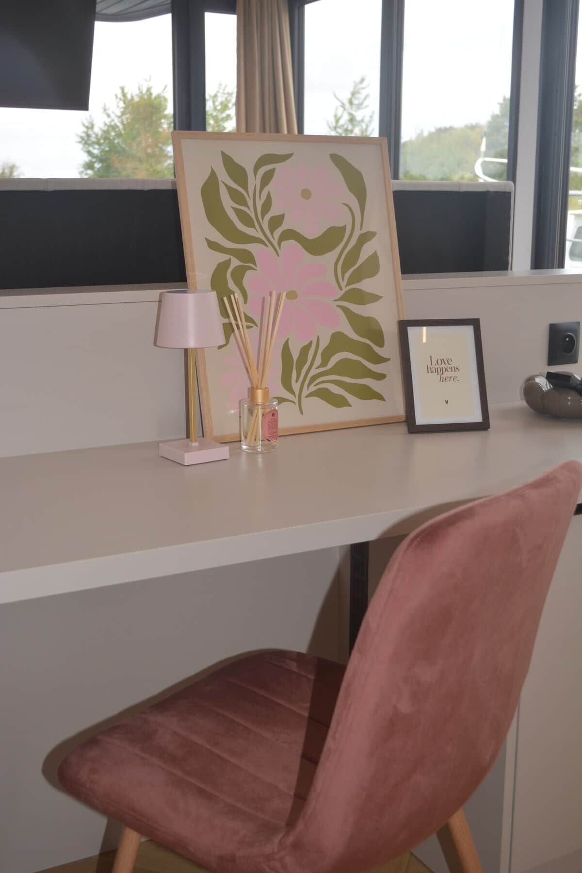Un espacio de trabajo con un escritorio, una silla rosa, una obra de arte enmarcada, una lámpara pequeña y un difusor de aroma de juncos.
