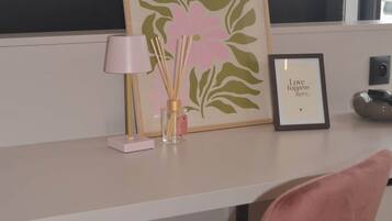 Un espacio de trabajo con un escritorio, una silla rosa, una obra de arte enmarcada, una lámpara pequeña y un difusor de aroma de juncos.